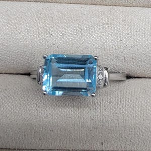 May include: A silver ring with a large, light blue, emerald-cut aquamarine gemstone. The ring is set with small, clear diamonds on the sides.