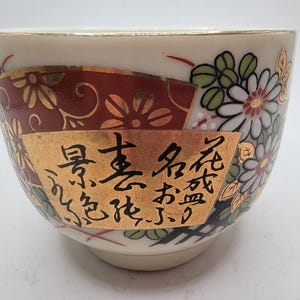 Puede incluir: Una taza de té de cerámica blanca con un borde dorado y diseños florales. La taza tiene un patrón floral rojo y dorado en un lado y un patrón floral blanco y negro en el otro lado. La taza también tiene una inscripción dorada en caracteres japoneses.