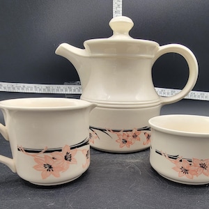 Puede incluir: Una tetera, un azucarero y una taza de té blancos con un diseño floral negro y rosa. La tetera tiene un asa y un pico. El azucarero y la taza de té tienen asas.