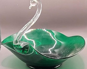 May include: A green glass swan-shaped bowl with a clear glass handle. The bowl is designed to look like a swan with its neck curved up and its head tilted to the side.
