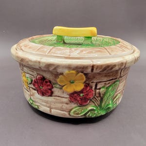 May include: A ceramic covered dish with a floral design. The dish is decorated with red, yellow, and green flowers on a brown background. The lid has a yellow handle.