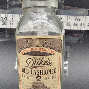 May include: A clear glass bottle with a black cap and a label that reads "Uncle Duke's Old Fashioned Spirit Drink". The label also features a skull and crossbones, a motorcycle, and the text "The Handmade Cocktail Co. Royal Tunbridge Wells".