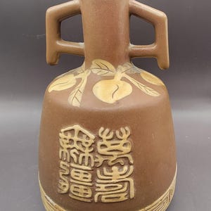 Puede incluir: Un jarrón de cerámica marrón con dos asas y un diseño de hojas y frutas. El jarrón tiene caracteres chinos en la parte delantera y un patrón geométrico alrededor de la base.