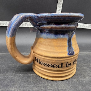 May include: Handmade ceramic mug with a tan base and a blue glazed rim and handle. The mug features the phrase "Blessed is th" in black lettering. The mug has a ribbed texture and a large handle.