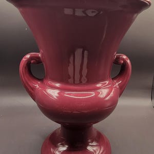 Puede incluir: Un jarrón de cerámica de color burdeos oscuro con dos asas y una base redonda. El jarrón tiene un acabado liso y brillante.