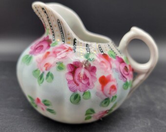 May include: A small white porcelain creamer with a floral design. The creamer has a handle and a spout. The floral design features pink roses and green leaves on a light blue background. The creamer is decorated with gold accents.