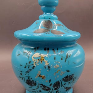 May include: A turquoise blue glass jar with a lid and a pointed finial. The jar is decorated with black floral designs.