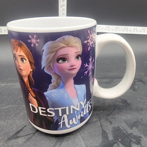 Puede incluir: Taza de cer&aacute;mica blanca con fondo morado que presenta un gr&aacute;fico de Elsa y Anna de la pel&iacute;cula Frozen. El texto "Destiny Awaits!" est&aacute; impreso en la taza.