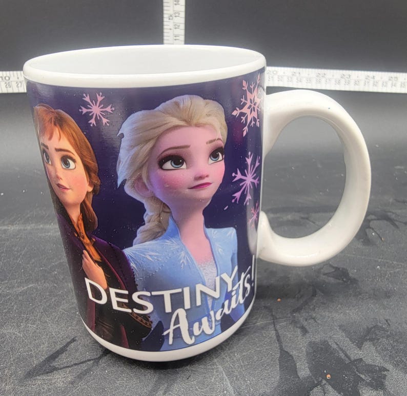 Puede incluir: Taza de cer&aacute;mica blanca con fondo morado que presenta un gr&aacute;fico de Elsa y Anna de la pel&iacute;cula Frozen. El texto "Destiny Awaits!" est&aacute; impreso en la taza.