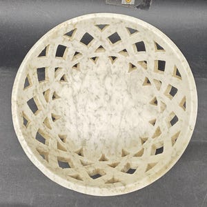 May include: A circular, off-white marble bowl featuring a geometric lattice design. The bowl has a shallow depth and a smooth interior. The lattice pattern comprises diamond and triangular cutouts, creating an openwork effect. Suitable for decorative or functional use.