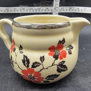 May include: A cream-colored ceramic pitcher with a silver rim and a floral design. The flowers are red with black leaves and stems.