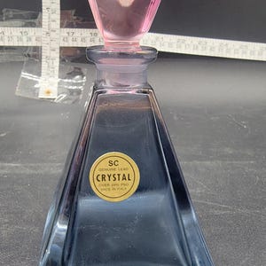 May include: A clear glass bottle with a pink glass stopper. The bottle is shaped like a pyramid and has a gold label that reads "SC Genuine Lead Crystal Over 24% PbO Made in Italy".