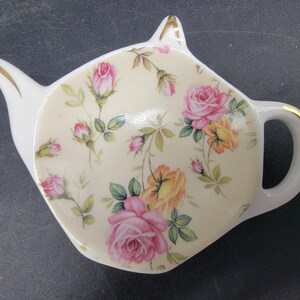 May include: A white ceramic teapot-shaped dish with a floral design. The dish is decorated with pink, yellow, and green roses and leaves on a cream background.