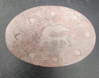 Può includere: Un sottobicchiere rotondo in pietra marrone rossiccia con un disegno scolpito di un elefante. L'elefante è scolpito in un colore grigio chiaro ed è circondato da un motivo di linee e punti.