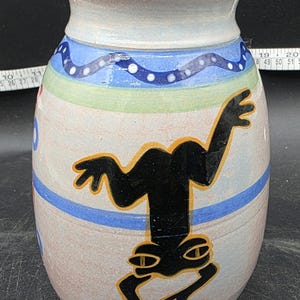 May include: A ceramic vase with a light beige base, adorned with horizontal bands of blue, green, and red. A black frog illustration with yellow accents is featured prominently on the vase. The vase has a wide body and a narrower neck.