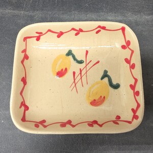 May include: A rectangular ceramic dish with rounded corners. The dish has a cream-colored base with a red border of stylized leaves. Two yellow and red fruit illustrations with green stems are painted on the dish.