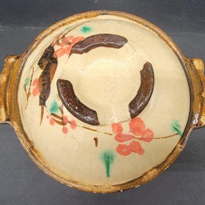 May include: A brown ceramic bowl with a lid, featuring a floral design in shades of pink, green, and brown. The bowl has a raised rim and a slightly rounded bottom.