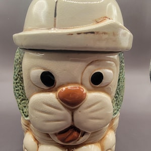 Peut inclure: Une boîte à biscuits en céramique en forme de chien portant un chapeau blanc. Le chien a des yeux bruns, un nez brun et une langue brune qui sort. Le chien porte une écharpe verte.