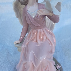 May include: A porcelain figurine of an angel with wings, wearing a pink dress and holding a white dove. The angel is sitting on a rock with flowers at the base.