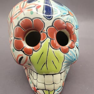 May include: A white ceramic skull with a colorful Day of the Dead design. The skull has red, green, blue, and black floral patterns. The eyes are black and the teeth are white.