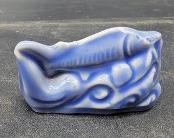 May include: A small, blue ceramic object with white accents. The object has a curved shape with a textured surface and a wave-like pattern. The ceramic piece appears to be a decorative item.