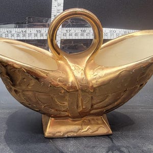 May include: A gold-colored, decorative serving dish with a handle. The dish has a square base and a textured surface. The interior is a cream color. The handle is a circular shape.