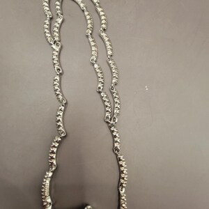 May include: A silver necklace with a wavy, textured design. The necklace is made of metal and has a shiny finish.