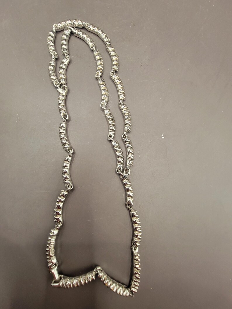 May include: A silver necklace with a wavy, textured design. The necklace is made of metal and has a shiny finish.