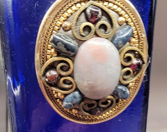 May include: A cobalt blue glass bottle with a decorative gold and silver metal cap. The cap features a white oval stone surrounded by smaller stones in shades of red, blue, and brown.