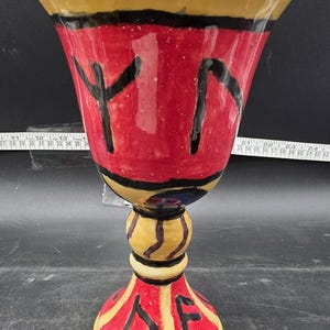 May include: A hand-painted ceramic goblet with a wide, red bowl and a yellow rim. The bowl features black abstract shapes. The stem is yellow with black stripes, and the base is red with yellow accents and black symbols.