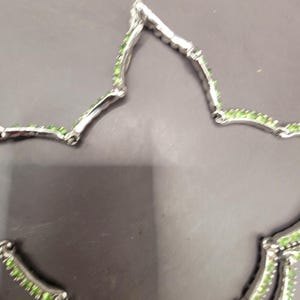 May include: A silver necklace with a delicate design featuring small green gemstones. The necklace is made of two strands of silver chain with a repeating pattern of small green stones.