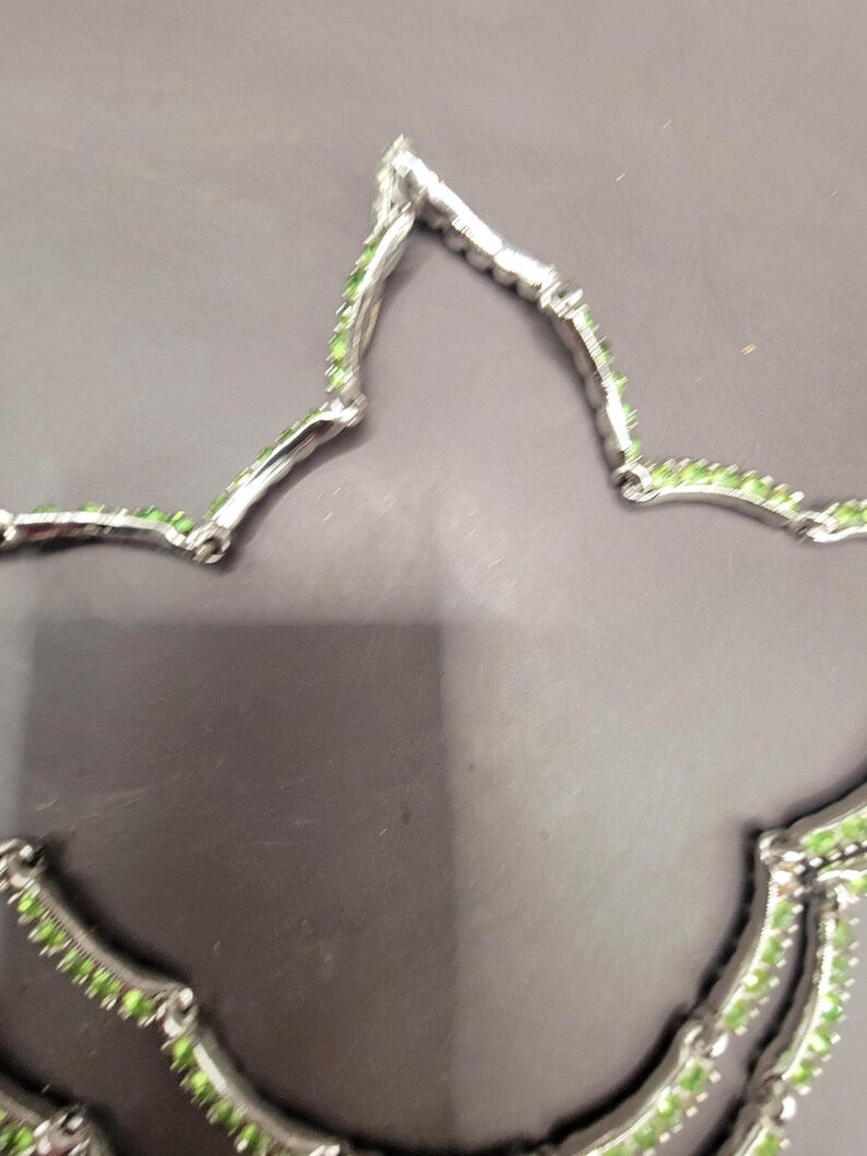 May include: A silver necklace with a delicate design featuring small green gemstones. The necklace is made of two strands of silver chain with a repeating pattern of small green stones.