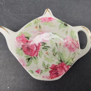 May include: White ceramic teapot-shaped tea bag holder with a pink and white floral design. The holder has a gold rim and a handle.