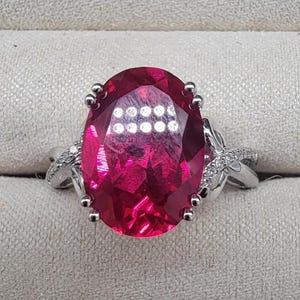 May include: A silver ring with a large, oval-shaped, red gemstone surrounded by smaller, clear gemstones.