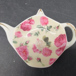 May include: White ceramic teapot-shaped dish with a pink rose floral pattern. The dish has a gold rim and a handle.