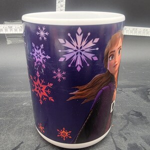 Puede incluir: Una taza de cer&aacute;mica blanca con fondo morado y un dise&ntilde;o que presenta a una ni&ntilde;a con cabello largo casta&ntilde;o, un vestido morado y copos de nieve en tonos de rosa, rojo y blanco.