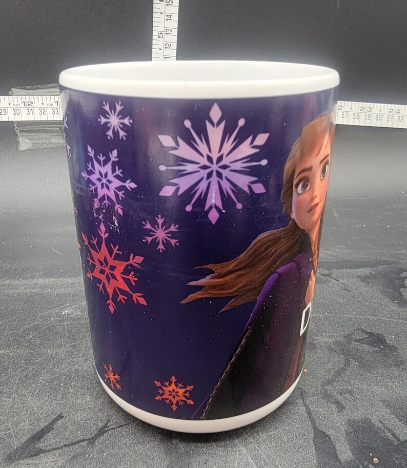 Puede incluir: Una taza de cer&aacute;mica blanca con fondo morado y un dise&ntilde;o que presenta a una ni&ntilde;a con cabello largo casta&ntilde;o, un vestido morado y copos de nieve en tonos de rosa, rojo y blanco.