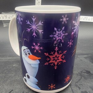Puede incluir: Una taza de cer&aacute;mica blanca con fondo azul y una imagen colorida de Olaf de la pel&iacute;cula Frozen. La taza est&aacute; decorada con copos de nieve en tonos rosa, morado y rojo.