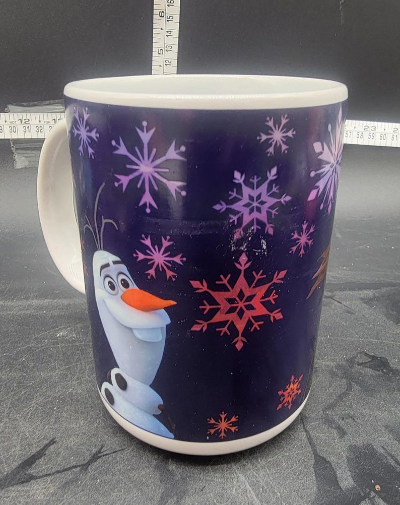 Puede incluir: Una taza de cer&aacute;mica blanca con fondo azul y una imagen colorida de Olaf de la pel&iacute;cula Frozen. La taza est&aacute; decorada con copos de nieve en tonos rosa, morado y rojo.