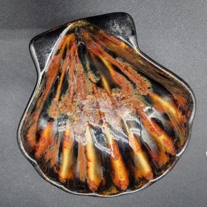 May include: A black ceramic seashell dish with a brown and orange glaze. The dish has a textured surface and is shaped like a scallop shell.
