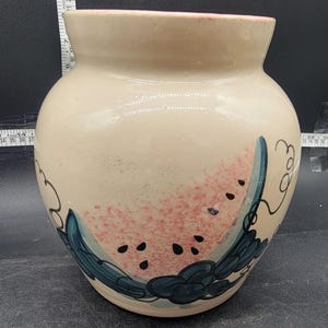 May include: A beige ceramic vase with a watermelon design. The vase features a pink watermelon slice with black seeds and blue-green leaves. The vase has a wide body and a slightly flared rim.