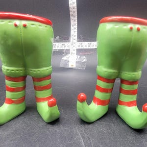 May include: Two green and red ceramic elf boot ornaments. The boots are striped with red and green and have a pointed toe. The boots are standing on a white surface.