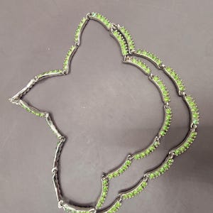 May include: A silver necklace with two rows of green stones. The necklace is made of small, round stones that are set in a silver setting. The necklace is a delicate and elegant piece of jewelry.