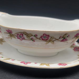 May include: A white porcelain gravy boat with a floral design in pink and green. The boat has a gold rim and sits on a matching saucer.