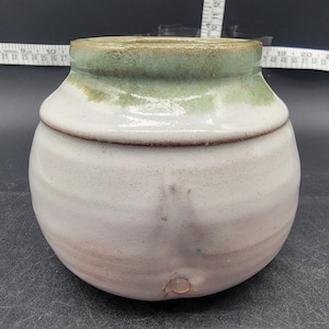May include: A white ceramic pot with a green glaze on the top rim. The pot has a rounded shape and a small hole in the bottom.