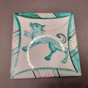 Op de afbeelding: Een vierkante keramische tegel met een witte achtergrond en een groen gestileerd dierontwerp. Het dier is een mythisch wezen met een lange staart en een lange snuit. De tegel heeft een groene rand met een geometrisch patroon.