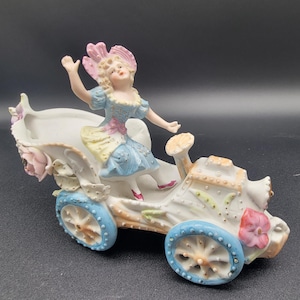 Może przedstawiać: Porcelanowa figurka przedstawiająca dziewczynkę w różowo-białym kapeluszu, niebieskiej sukience i białych butach, jadącą w biało-niebieskim powozie z kwiatowymi akcentami.