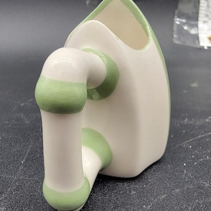 Puede incluir: Una taza de cerámica con forma de plancha vintage, con un cuerpo blanco y un asa y ribetes verdes. La taza tiene una parte superior triangular y un asa curvada, diseñada para parecerse a una plancha antigua.