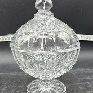 May include: A clear, cut-glass candy dish with a lid. The dish has a round bowl shape, a pedestal base, and a decorative finial on the lid. The glass has a diamond and fan pattern. The dish is approximately 8 inches tall.