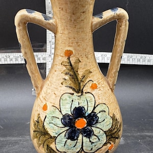 May include: A tan ceramic vase with two handles and a dark blue rim. The vase features a large floral design with blue and green petals and an orange center. Orange dots and green leaves accent the design.